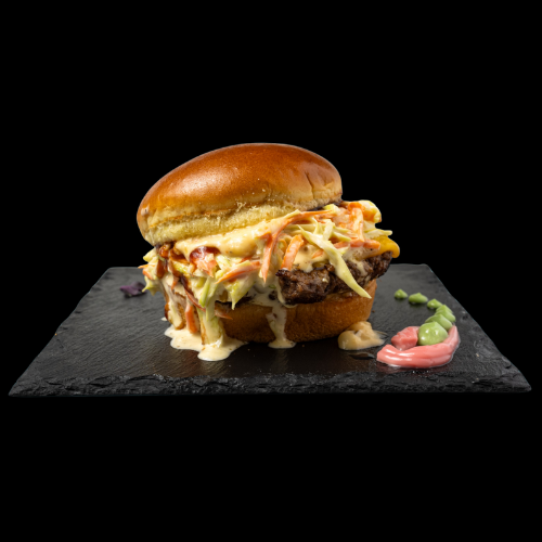 Ein saftiger Burger mit cremigem Krautsalat und Soße, serviert auf einem glänzenden Brioche-Bun, liegt auf einer schwarzen Schieferplatte mit grünen und rosa Soßen an der Seite.