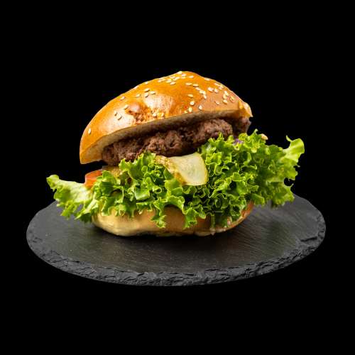 Ein Cheeseburger mit Salat, Tomate, Käse und einem Rinderpatty in einem Sesambrötchen, serviert auf einer runden schwarzen Schieferplatte vor schwarzem Hintergrund.