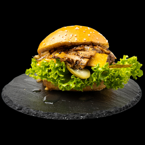 Ein Gourmet-Burger mit Sesambrötchen, grünem Blattsalat, geschmolzenem Käse, sautierten Champignons, Essiggurken und einer Rindfleischpastete, serviert auf einer runden schwarzen Schieferplatte vor einem schwarzen Hintergrund.