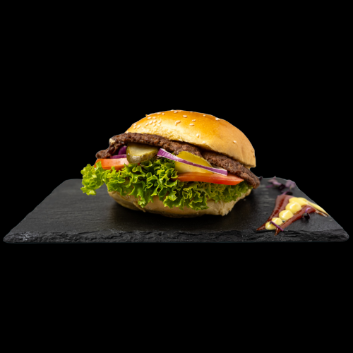 Ein Rindfleisch-Burger mit Salat, Tomaten, Essiggurken, roten Zwiebeln und Käse in einem Sesambrötchen, serviert auf einer schwarzen Schieferplatte mit einer kleinen Beilage an der Seite, vor einem schwarzen Hintergrund.