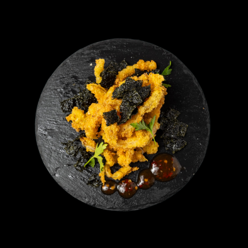 Eine schwarze Schieferplatte mit knusprig goldgelb gebratenen Calamari, garniert mit dunklen Seetangstücken, frischen Kräutern und drei Klecksen dunkler Dip-Sauce an der Seite.