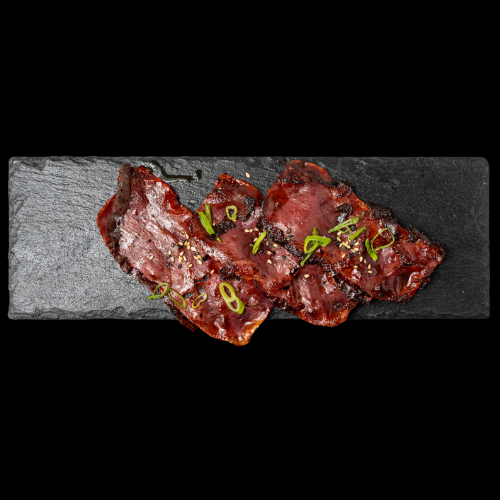 Scheiben von glasiertem gegrilltem Fleisch, garniert mit gehackten grünen Zwiebeln, serviert auf einer rechteckigen schwarzen Schieferplatte vor weißem Hintergrund.