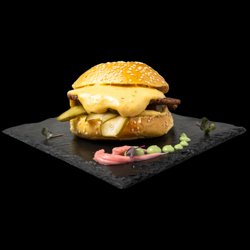 Ein Cheeseburger mit Essiggurken und einem Sesambrötchen liegt auf einem Schieferbrett, garniert mit rosa und grünen Saucen und kleinem Grünzeug, auf einem schwarzen Hintergrund.