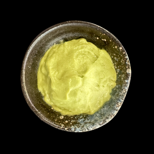 Eine kleine Keramikschale, gefüllt mit cremig-grüner Guacamole, fotografiert vor einem schwarzen Hintergrund.