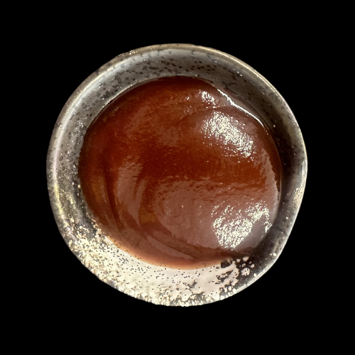 Eine kleine Keramikschüssel, gefüllt mit einer dicken, glänzenden, rotbraunen Sauce, auf schwarzem Hintergrund.