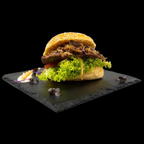 Ein Gourmet-Burger mit Salat, Tomate, gegrillten Zwiebeln und einem Sesambrötchen wird auf einer schwarzen Schieferplatte serviert, garniert mit Sauce und essbaren Blumen vor einem schwarzen Hintergrund.