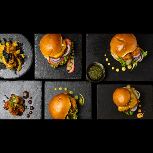 Sechs schwarze quadratische Teller in zwei Reihen, auf denen jeweils Gourmet-Burger mit verschiedenen Belägen und Garnierungen liegen; auf einem Teller befinden sich gebratene Vorspeisen und auf einem anderen eine kleine Schale mit grüner Soße. Der Hintergrund ist schwarz.