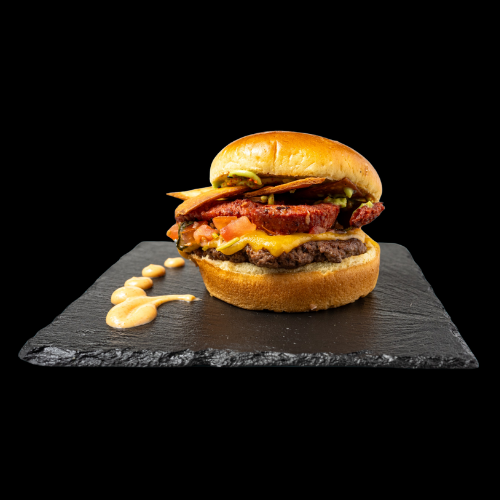 Ein Gourmet-Burger mit Speck, Cheddar-Käse, Tomate, Salat und Soße, serviert auf einer schwarzen Schiefertafel mit einem Strudel aus cremiger Soße daneben, alles vor einem schwarzen Hintergrund.