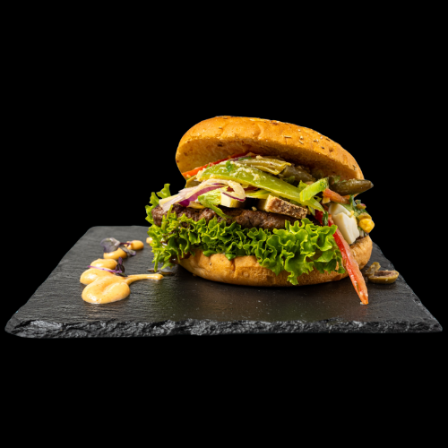 Ein Gourmet-Veggie-Burger mit Salat, Tomaten, Essiggurken, Zwiebeln und Soße auf einem entkernten Brötchen, serviert auf einer schwarzen Schieferplatte mit Soßentupfern an der Seite, vor einem schwarzen Hintergrund.